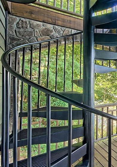 Unique outdoor spiral staircase with black aluminum treads and railing, installed on a lakeside deck with stone columns and scenic water views.