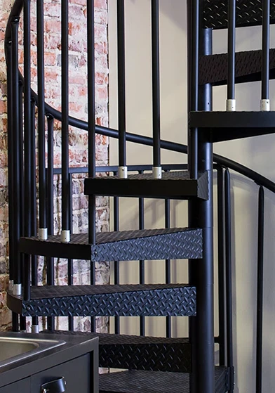 Black powder-coated spiral staircase with diamond plate treads, installed in an urban interior with exposed brick walls.