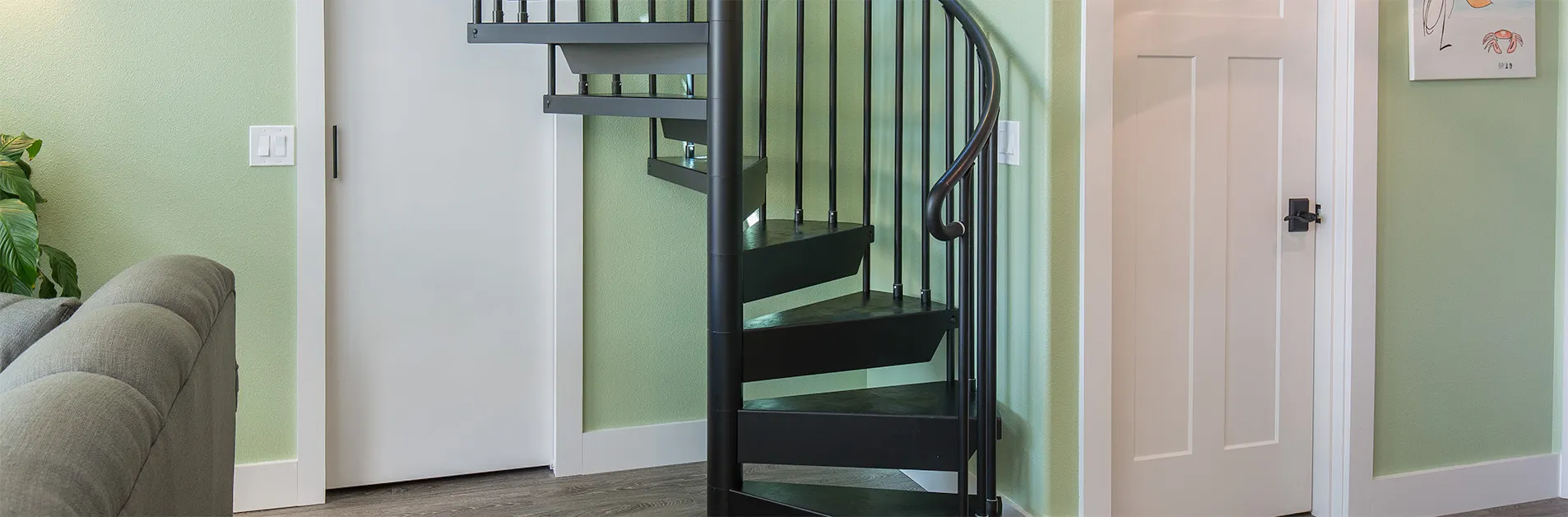 Compact black steel spiral staircase installed in a small living space beside doors and sofa, showing its minimal footprint.