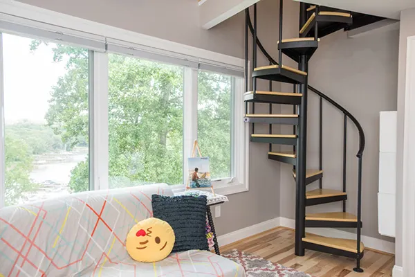 Compact black steel spiral staircase with light wood treads leading to a loft mezzanine beside large windows, showcasing space-saving modern staircase design.