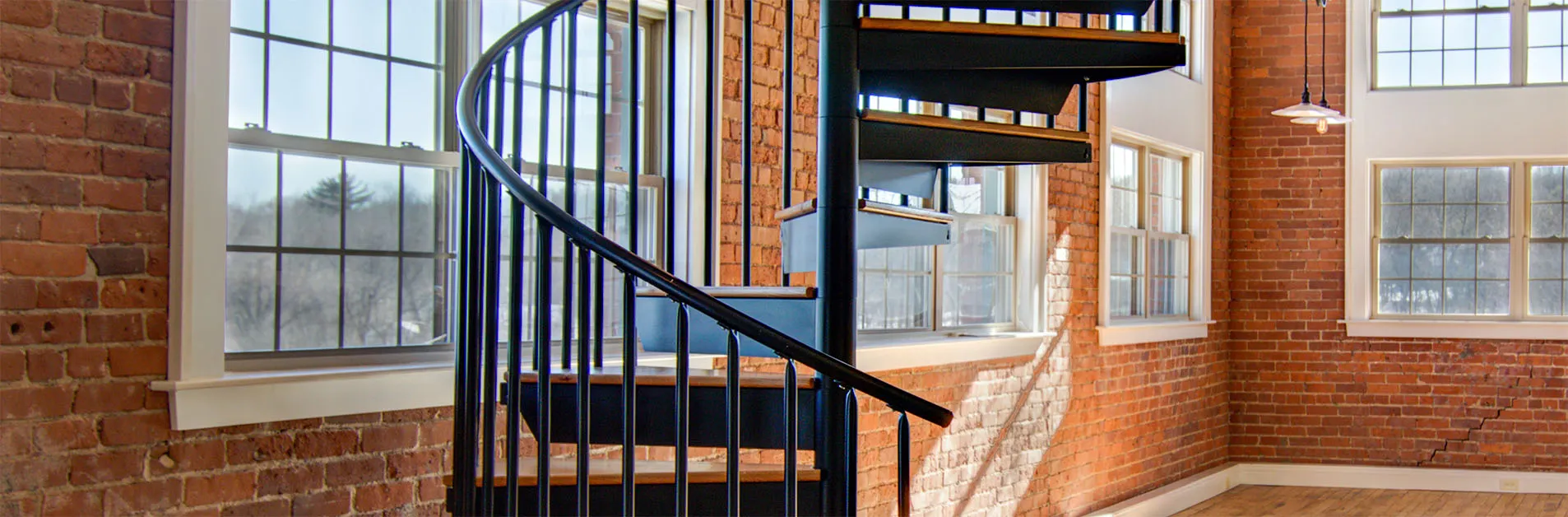 Black steel spiral staircase with wood treads set against exposed brick walls and tall windows, emphasizing industrial design and architectural character.