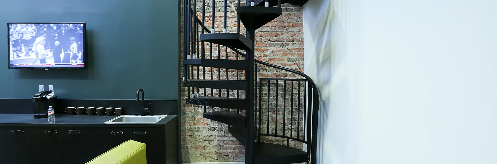 Black steel spiral staircase in an urban loft with exposed brick wall and kitchenette.