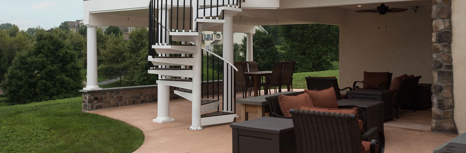White outdoor spiral staircase connecting an upper deck to a covered patio with outdoor seating and a stone column base.