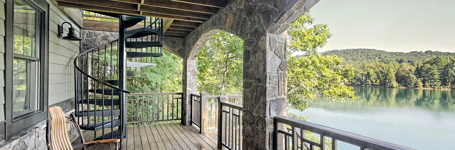 Black galvanized steel spiral staircase connecting a covered deck overlooking a scenic lake and forested hills.